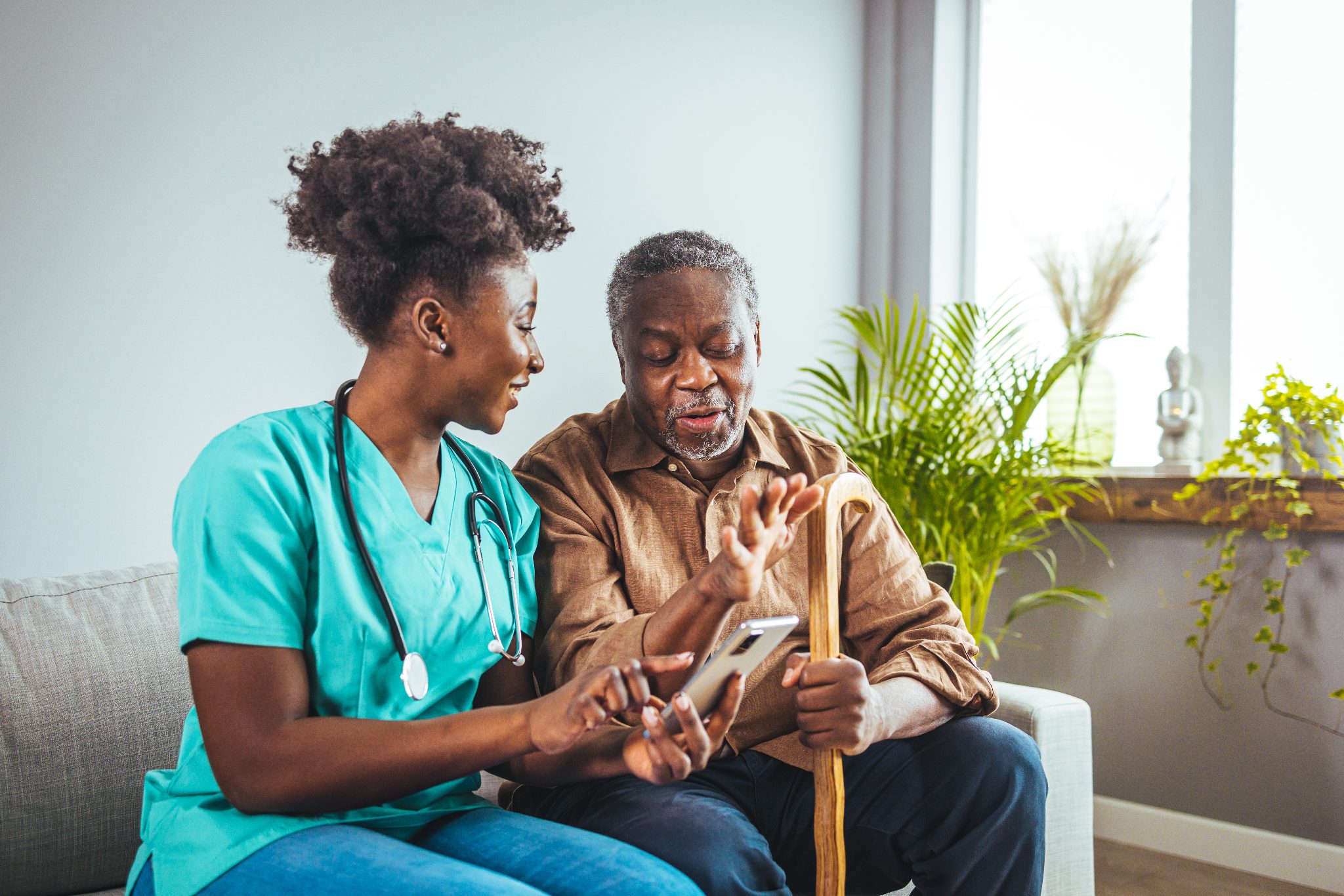 Healthcare worker in teal scrubs discussing smartphone with elderly male patient holding cane